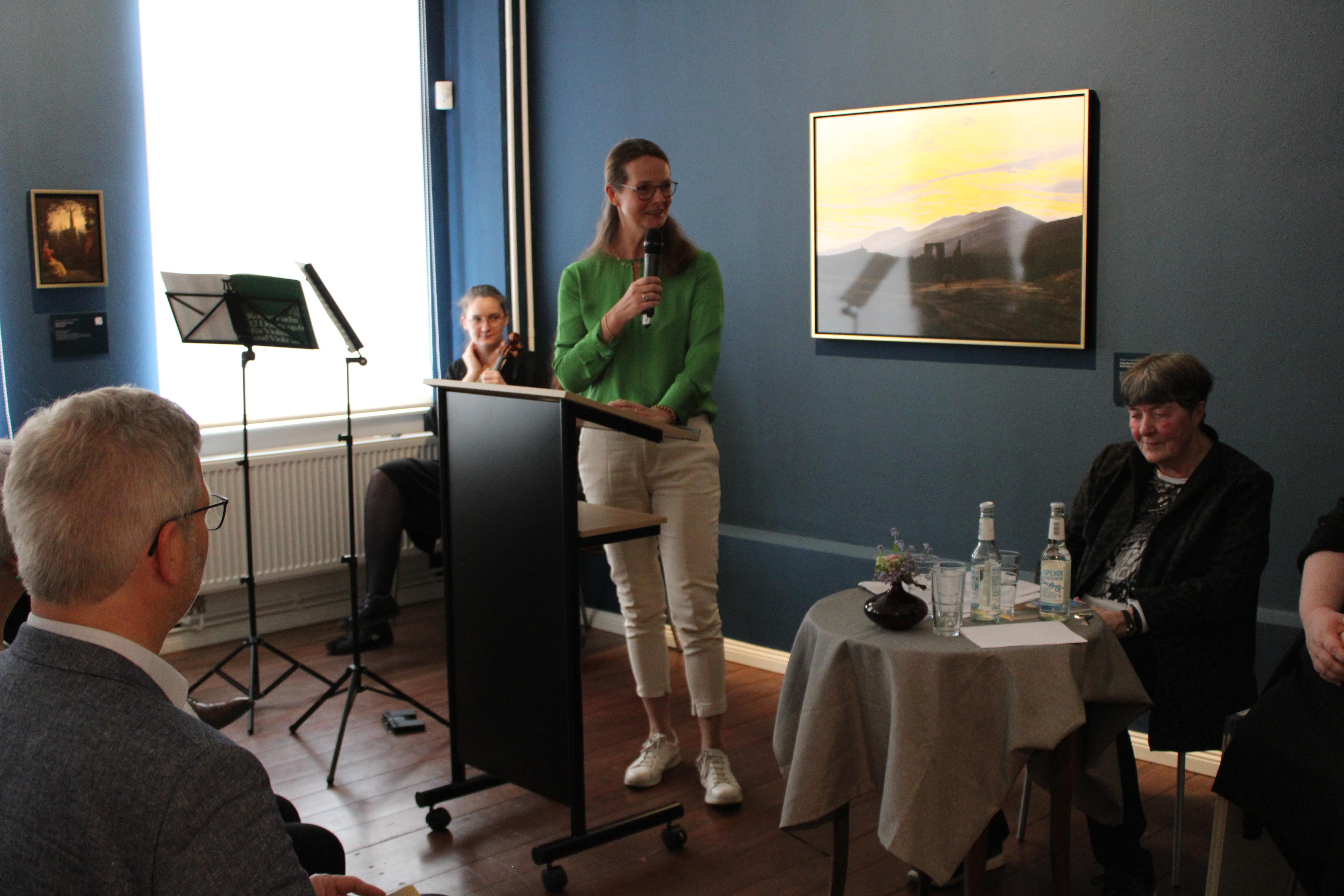 Eine Frau, Bettina Martin, hält eine Rede. Eine weitere Frau, Hannelore Kohl, sitzt im Hintergrund auf einem Stuhl auf der Bühne.